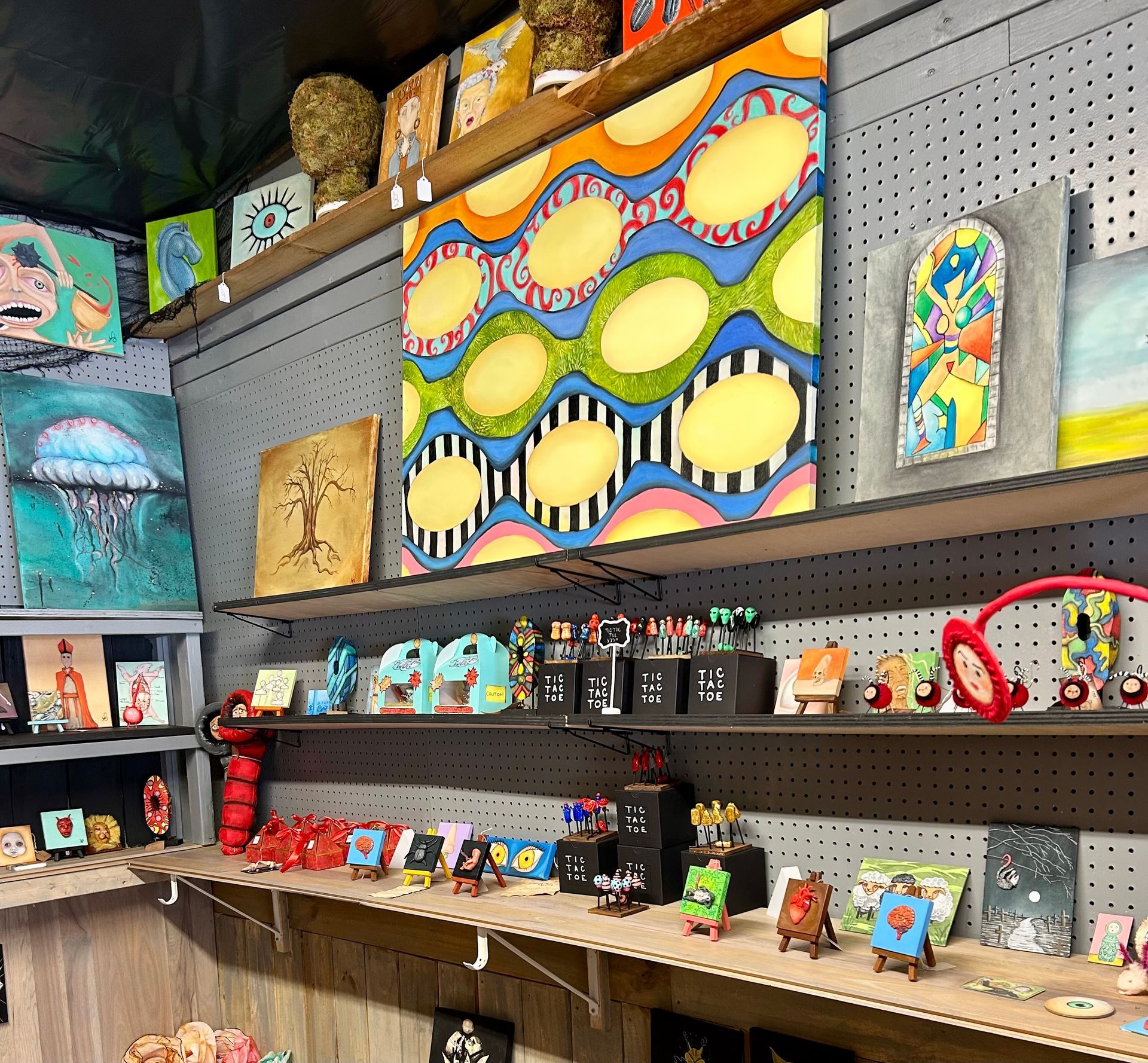 A shop wall featuring diverse colorful paintings, artwork, and small sculptures displayed on shelves and pegboards.