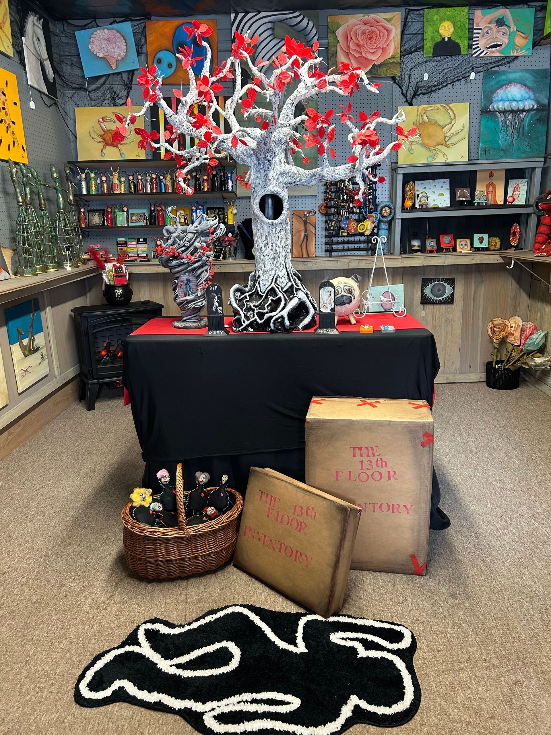 A room with a central black-covered table, a white tree sculpture with red leaves, floor bags, and a body outline rug.