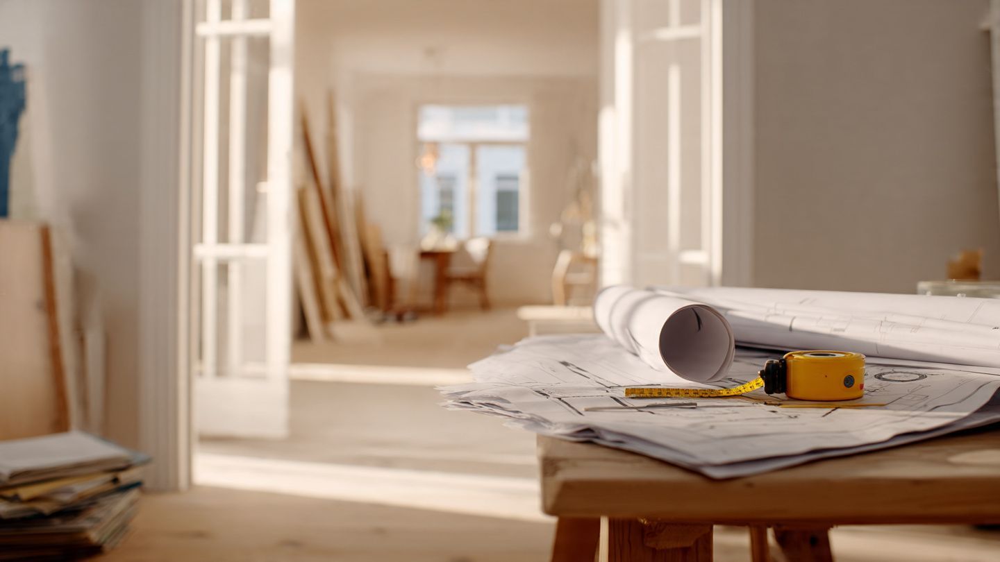 A tape measure and blueprints rest on a wooden table in a bright, sunlit room undergoing construction.