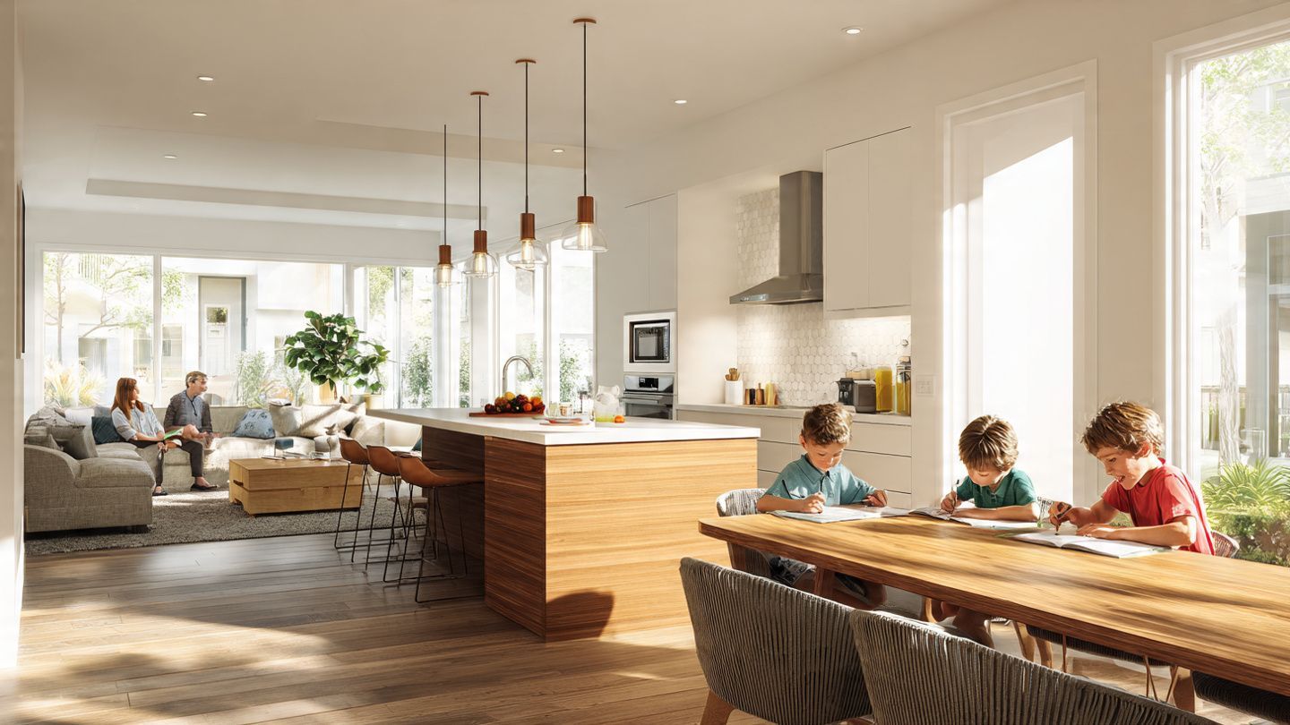 A bright, open-concept kitchen and living area with people socializing and children seated at a long wooden table.