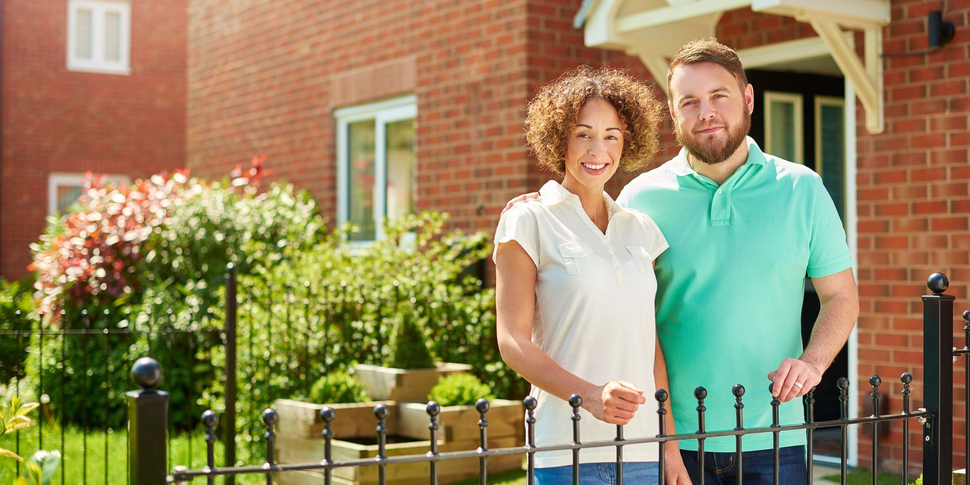 Couple standing in front of a brick house, smiling. They are near a fence, with greenery visible.