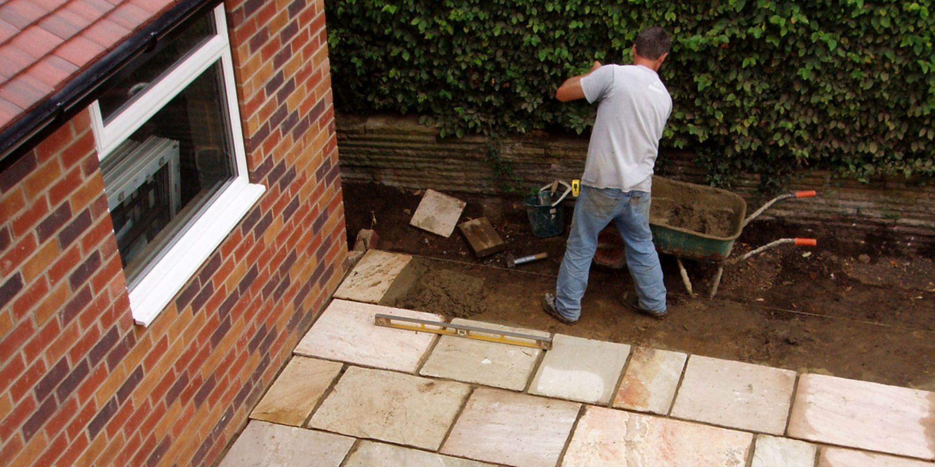 Man laying patio stones next to a brick building and a hedge.