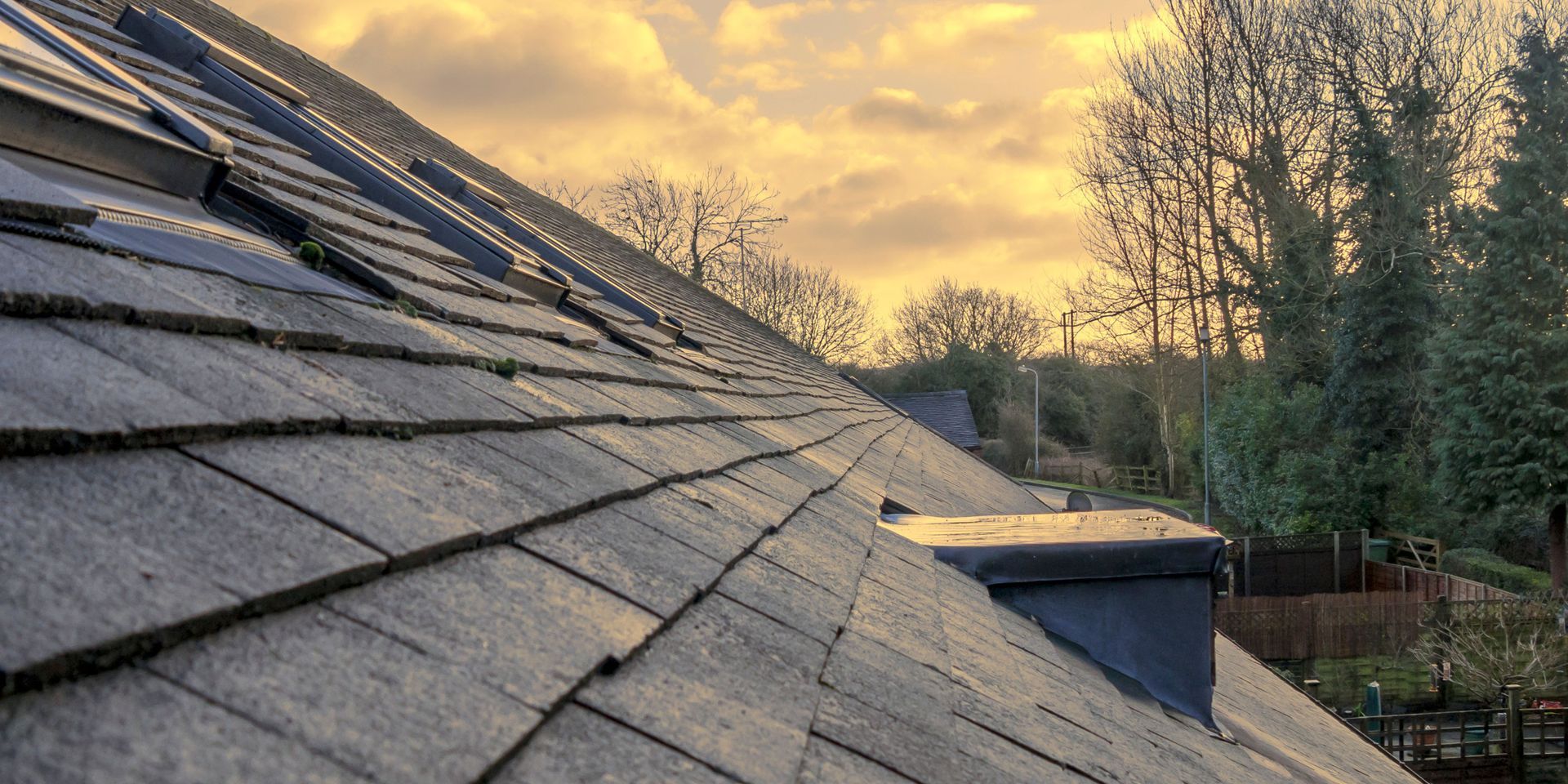Slate roof on a house, with a chimney in the foreground. Trees and a yellow sky in the background.