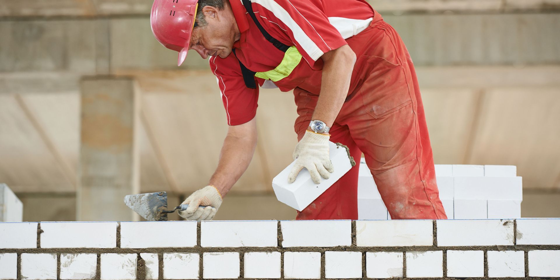 Construction worker in red overalls laying white bricks, wearing a hard hat and gloves.
