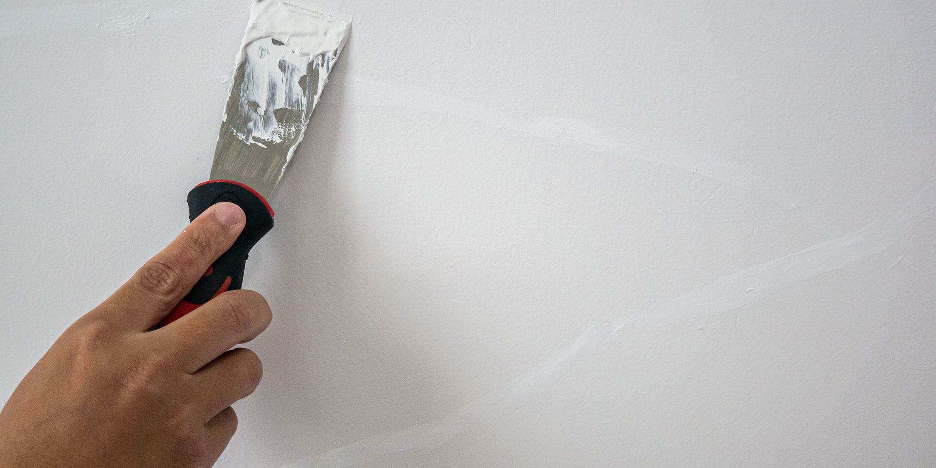 Hand using a putty knife to apply spackle to a white wall.