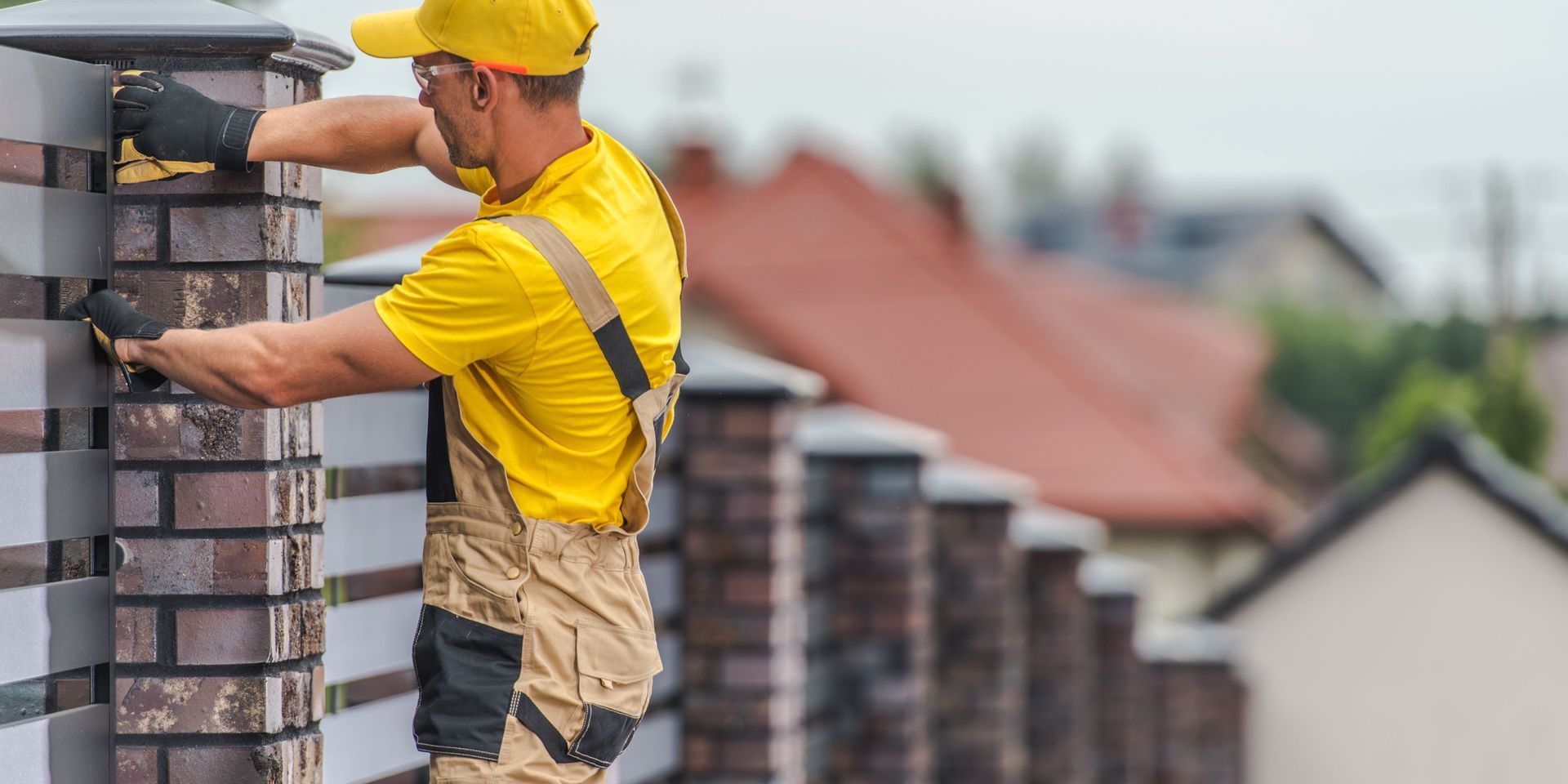 Man in yellow shirt and overalls working on a brick fence, wearing gloves and a yellow hat.