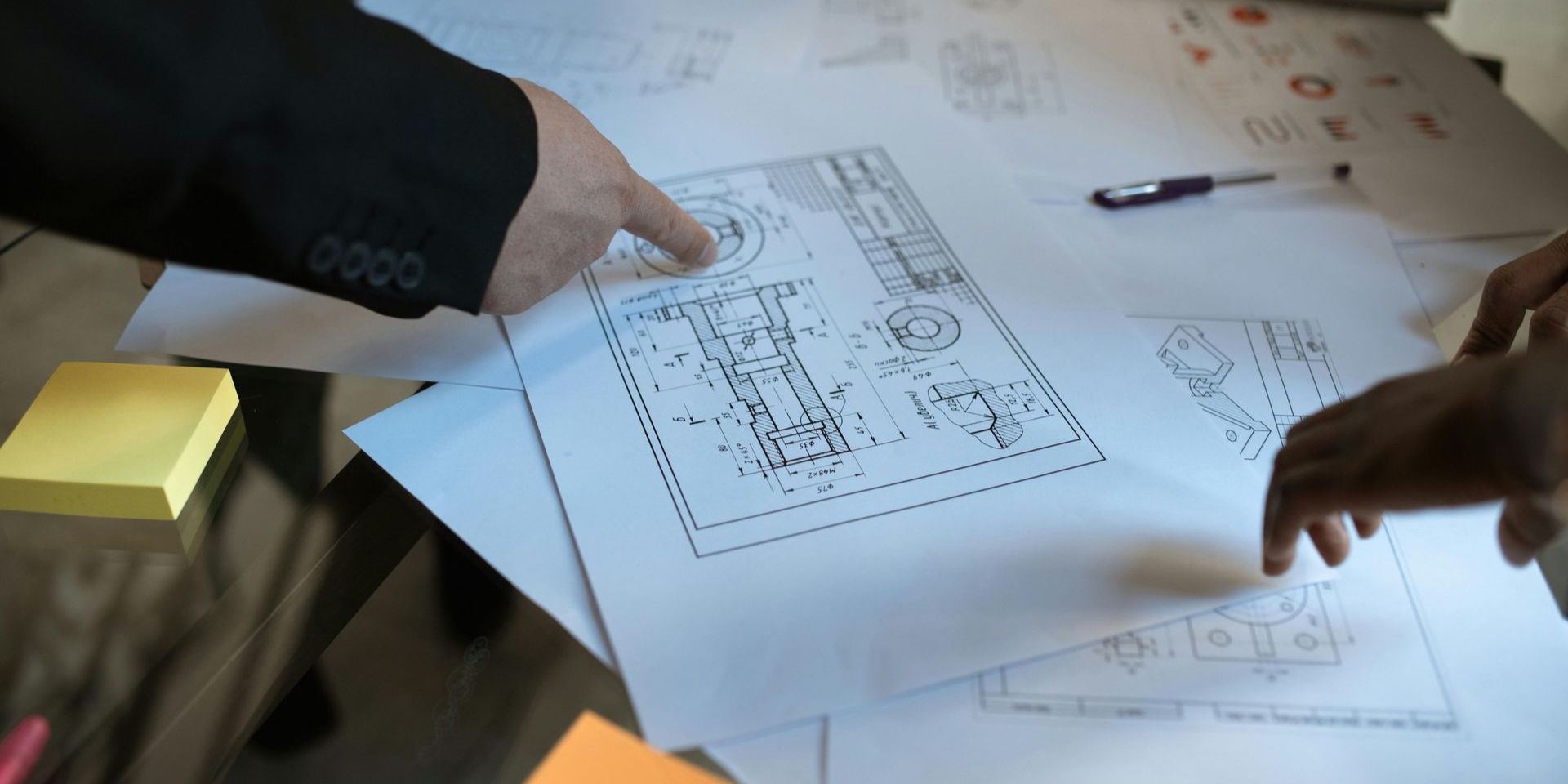 Two people's hands pointing at blueprints on a table. One person wears a black suit, and post-it notes are on the left.