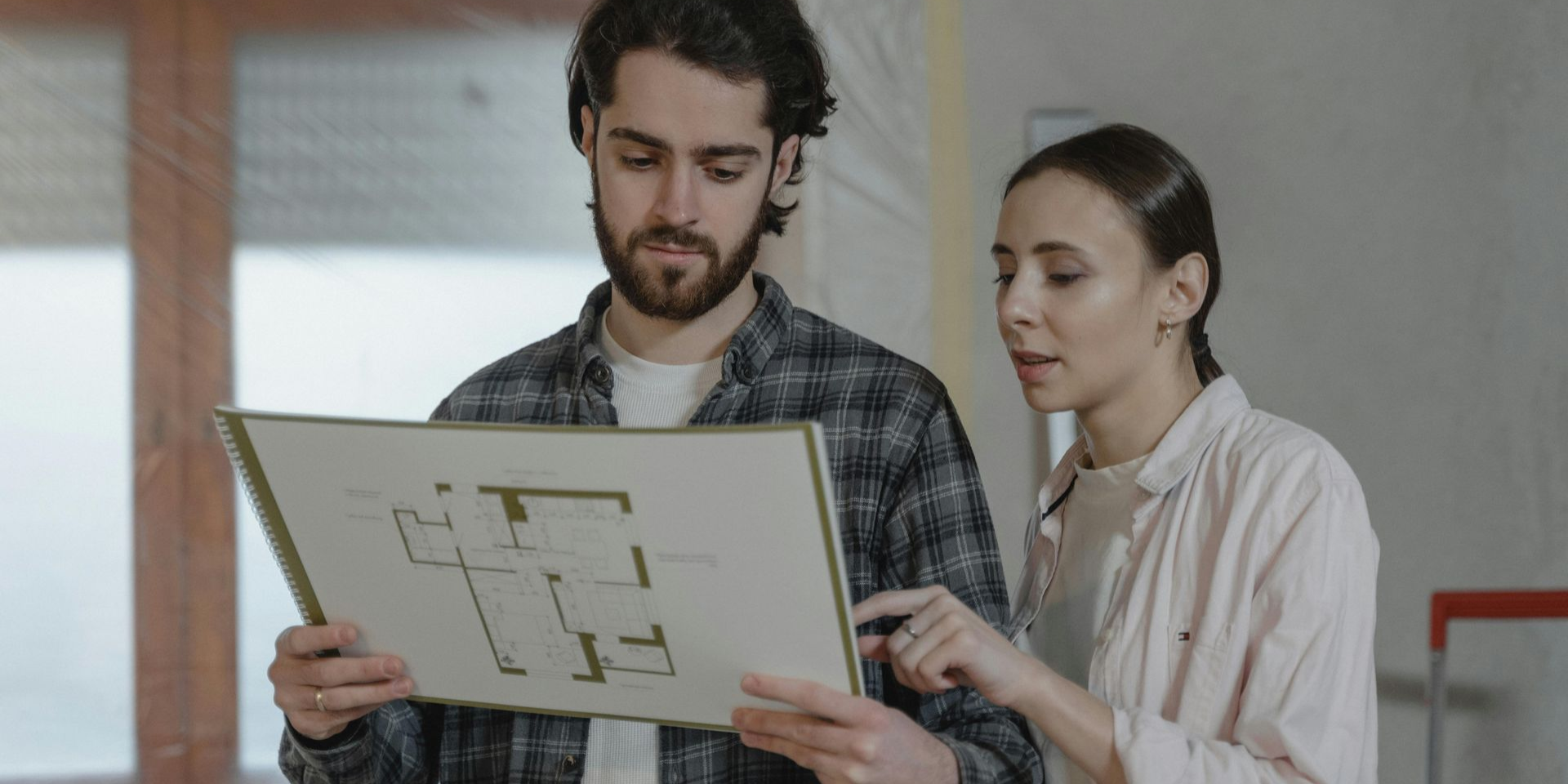 A couple reviews blueprints in a room with a window, discussing a floor plan.