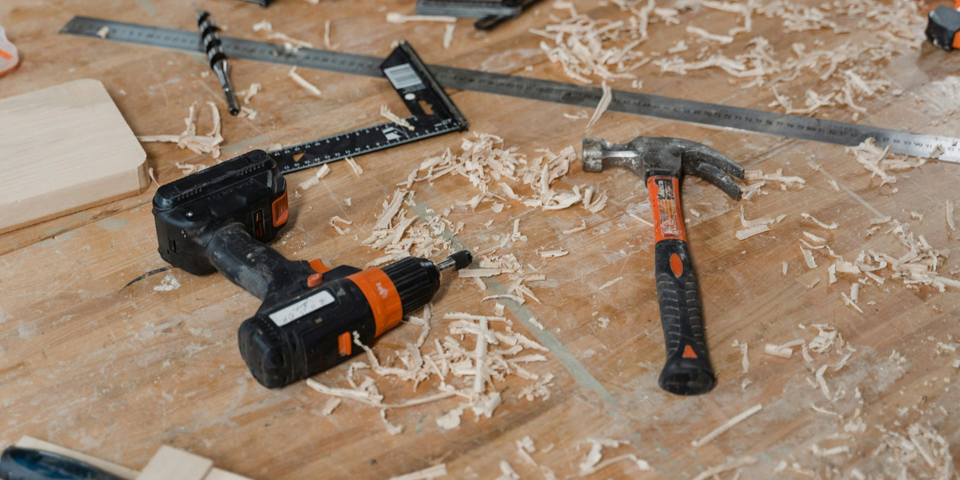 Woodworking tools on a wooden surface: drill, hammer, ruler, square, and scattered wood shavings.