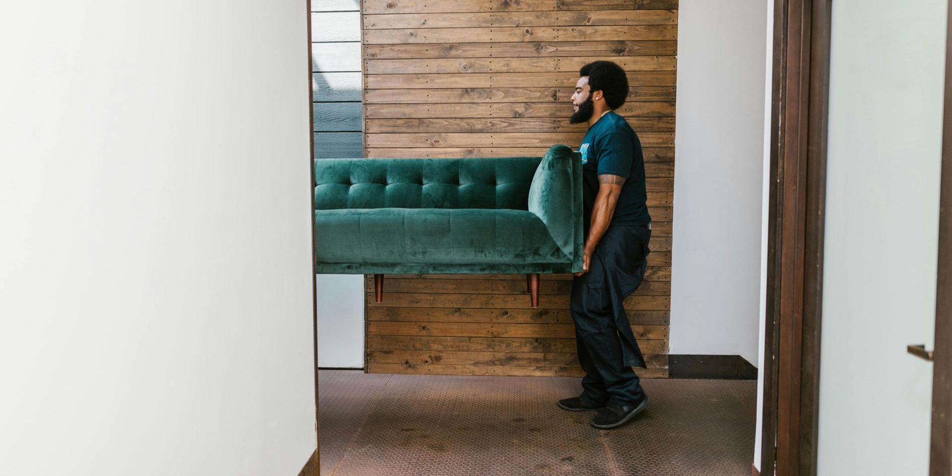 A person carrying a teal couch through a doorway. The walls are white and wood-paneled.