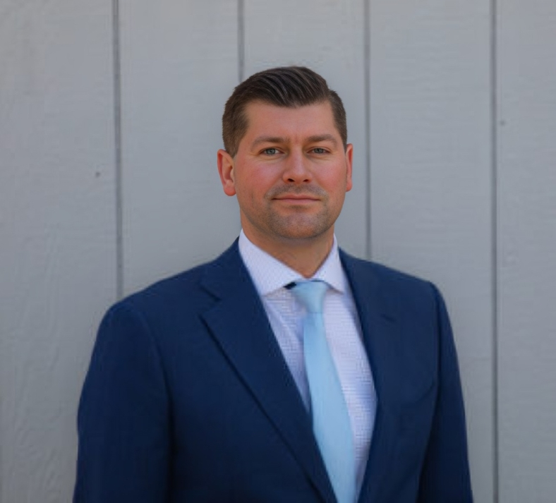 Man in a blue suit and light blue tie against a gray wall, smiling.