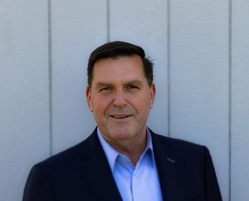Man in blue shirt and navy blazer smiles in front of a gray wall.