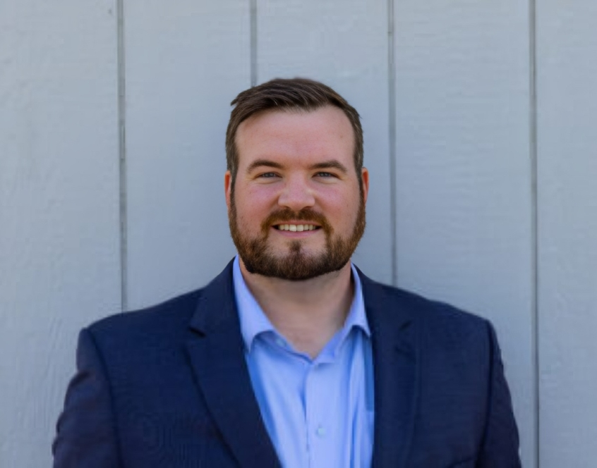 Man in a blue blazer and button-up shirt smiling in front of a gray wall.