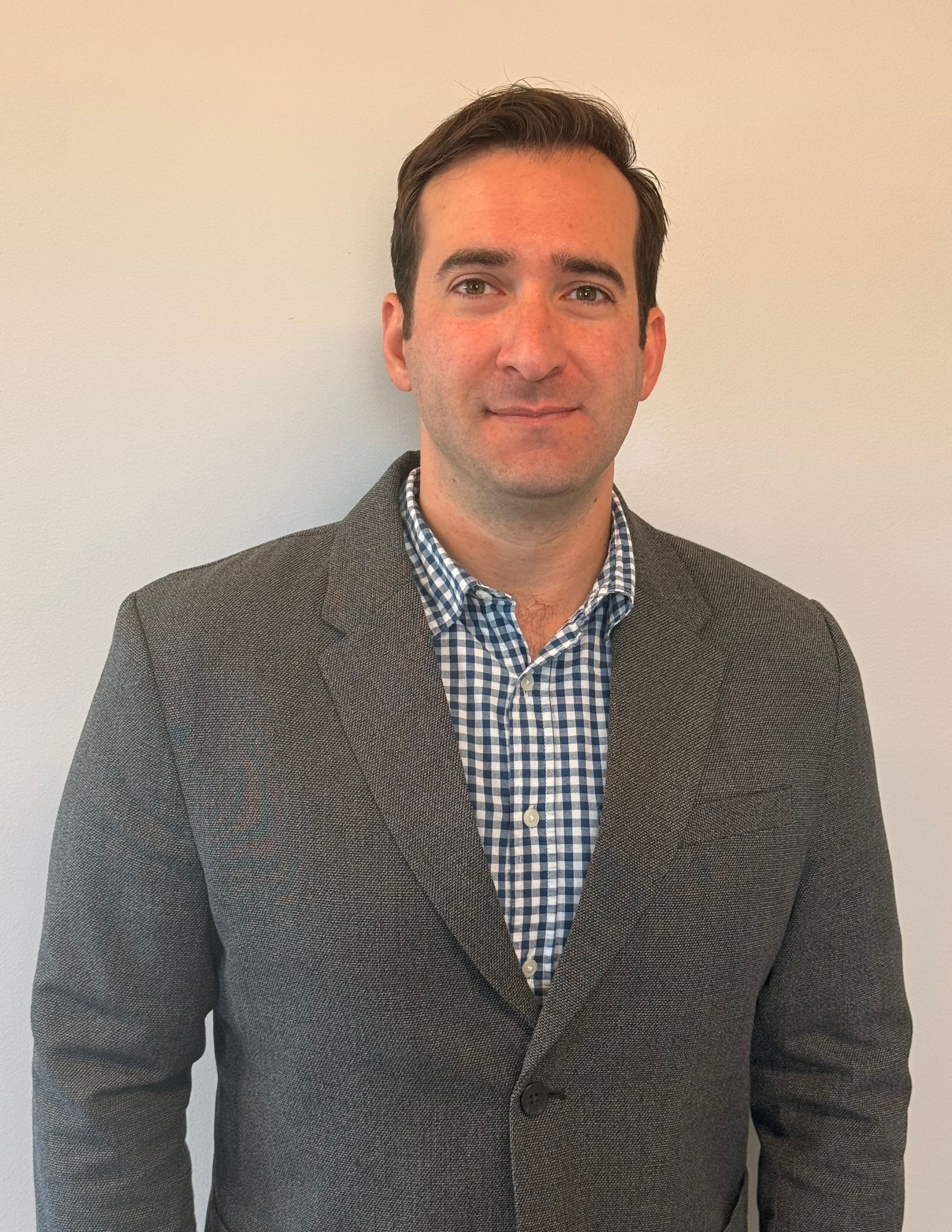 Man in blazer and checkered shirt, standing against a plain wall.