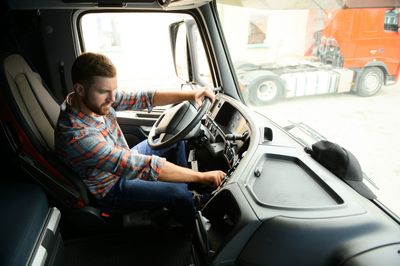 Driver in cab of truck turning ignition. Another truck visible through window.