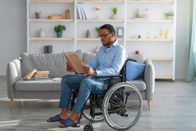 Man in wheelchair reading papers in a living room, seated in front of a gray couch.