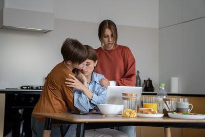 Three people comfort each other at a kitchen table, looking at a laptop. One is hugging another who looks sad.