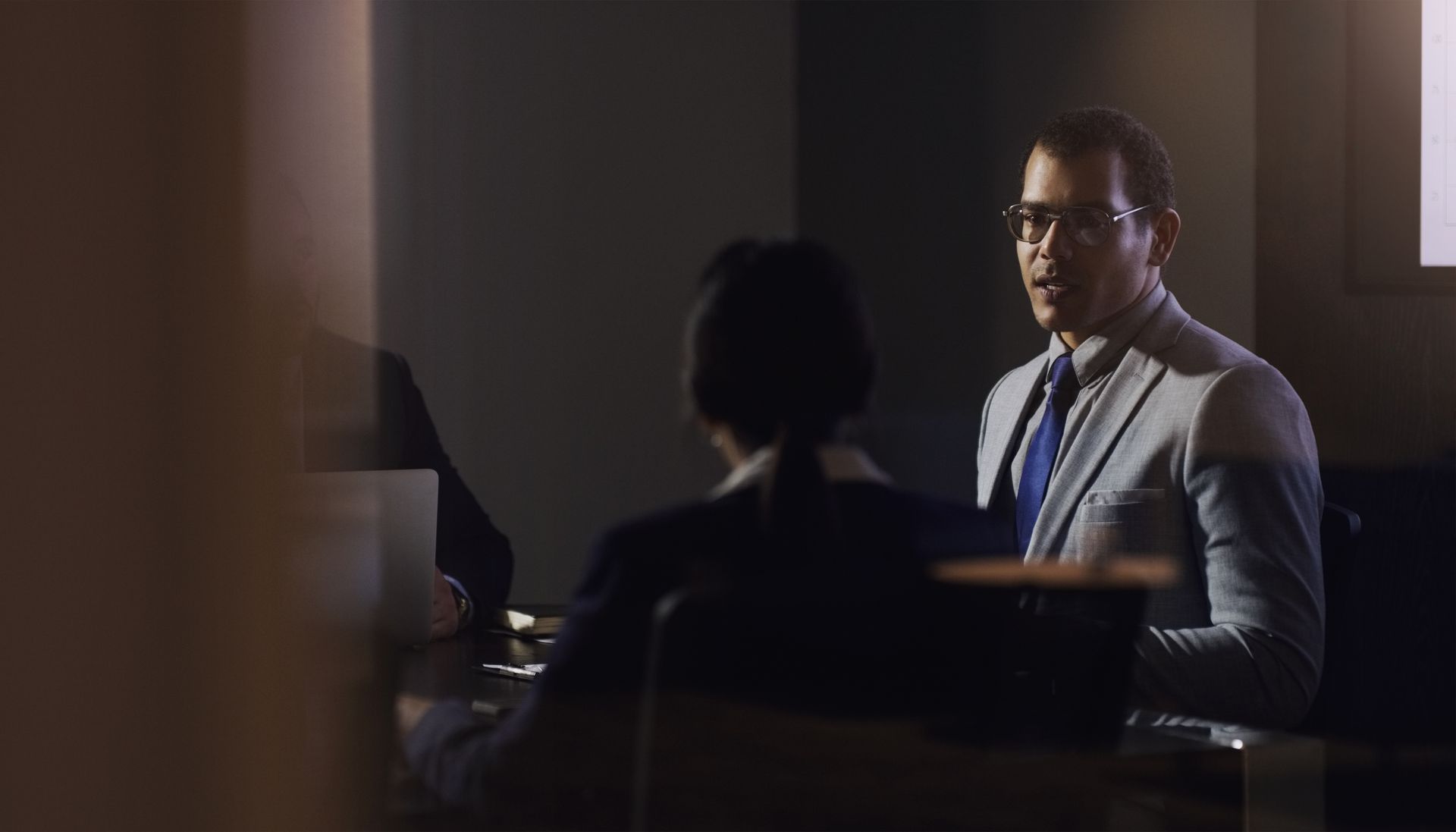 Man in glasses, gray suit, blue tie, speaking to person seated at a table. Dark office setting.