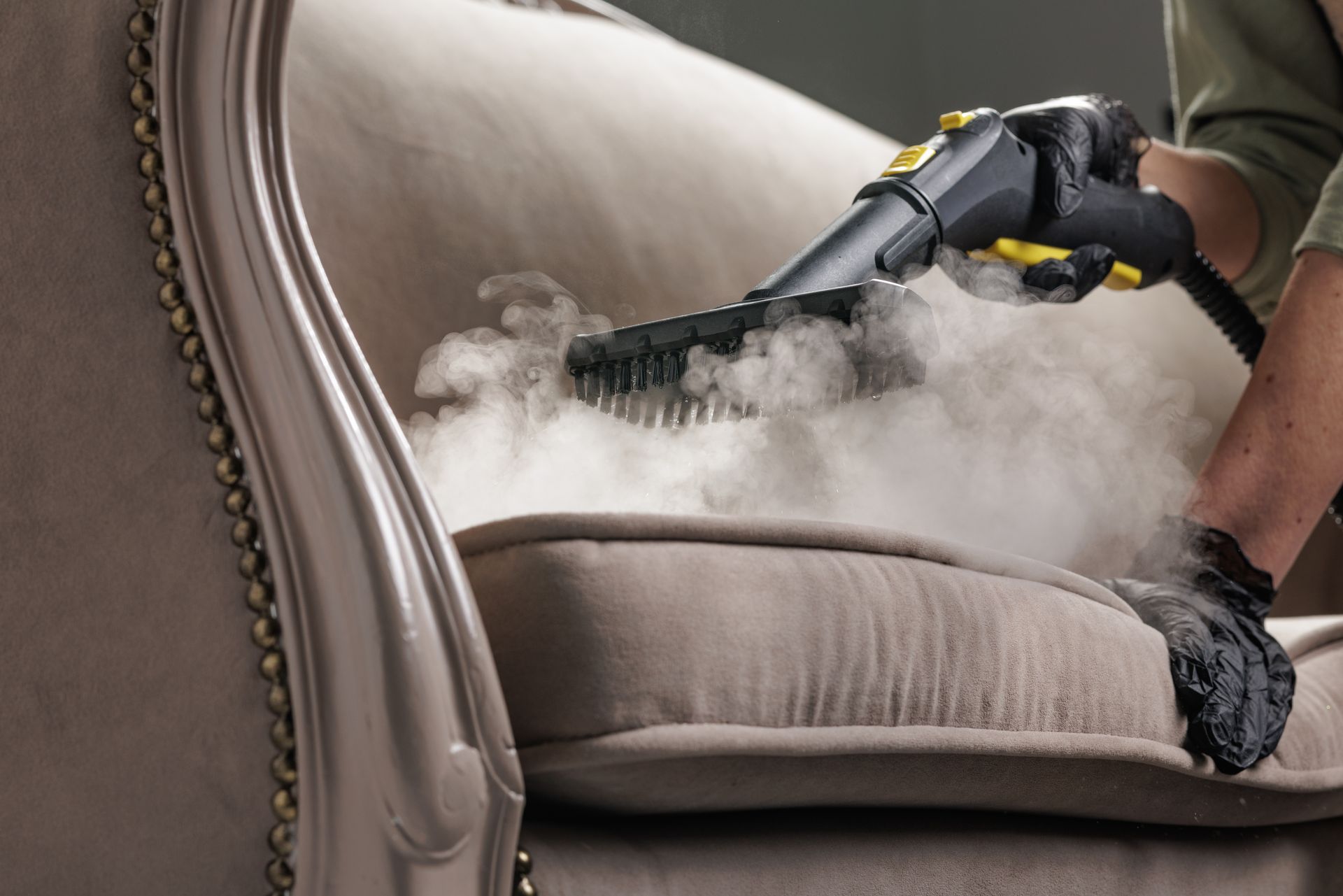 A person in black gloves uses a handheld steam cleaner on a light brown upholstered chair.