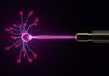 A bright purple electrical discharge from a plasma ball connecting to a metallic sensor or probe against a black backdrop.