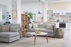 A bright, modern open-plan apartment with gray sofas, a wood bookshelf, a round coffee table, and a kitchen in the back.