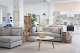 A bright, modern open-plan apartment with gray sofas, a wood bookshelf, a round coffee table, and a kitchen in the back.