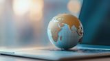 A small globe with textured gold continents resting on the keyboard of a laptop in a softly blurred, neutral setting.