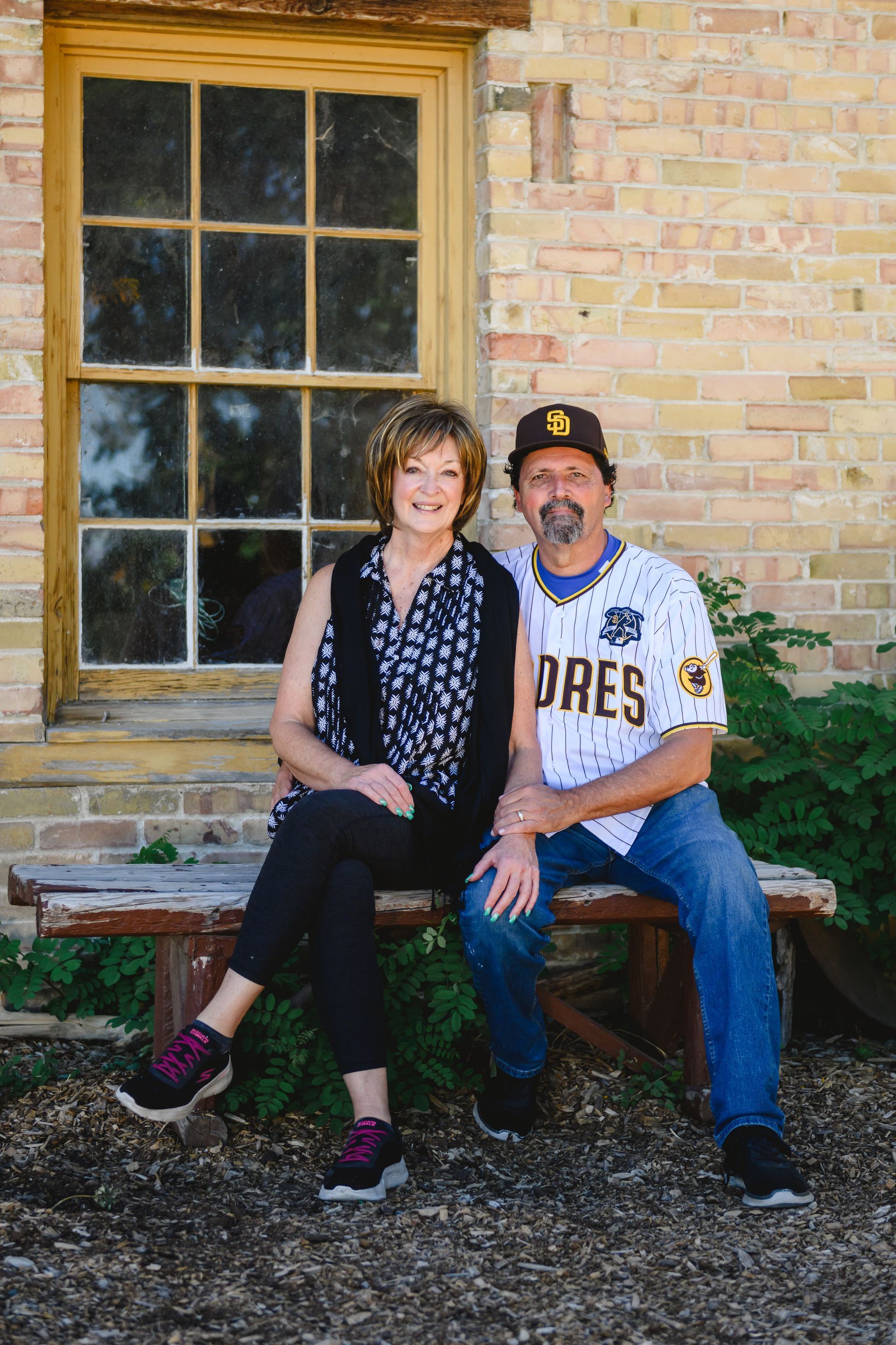 A couple sitting on a rustic bench in front of a brick building with a large pane window.