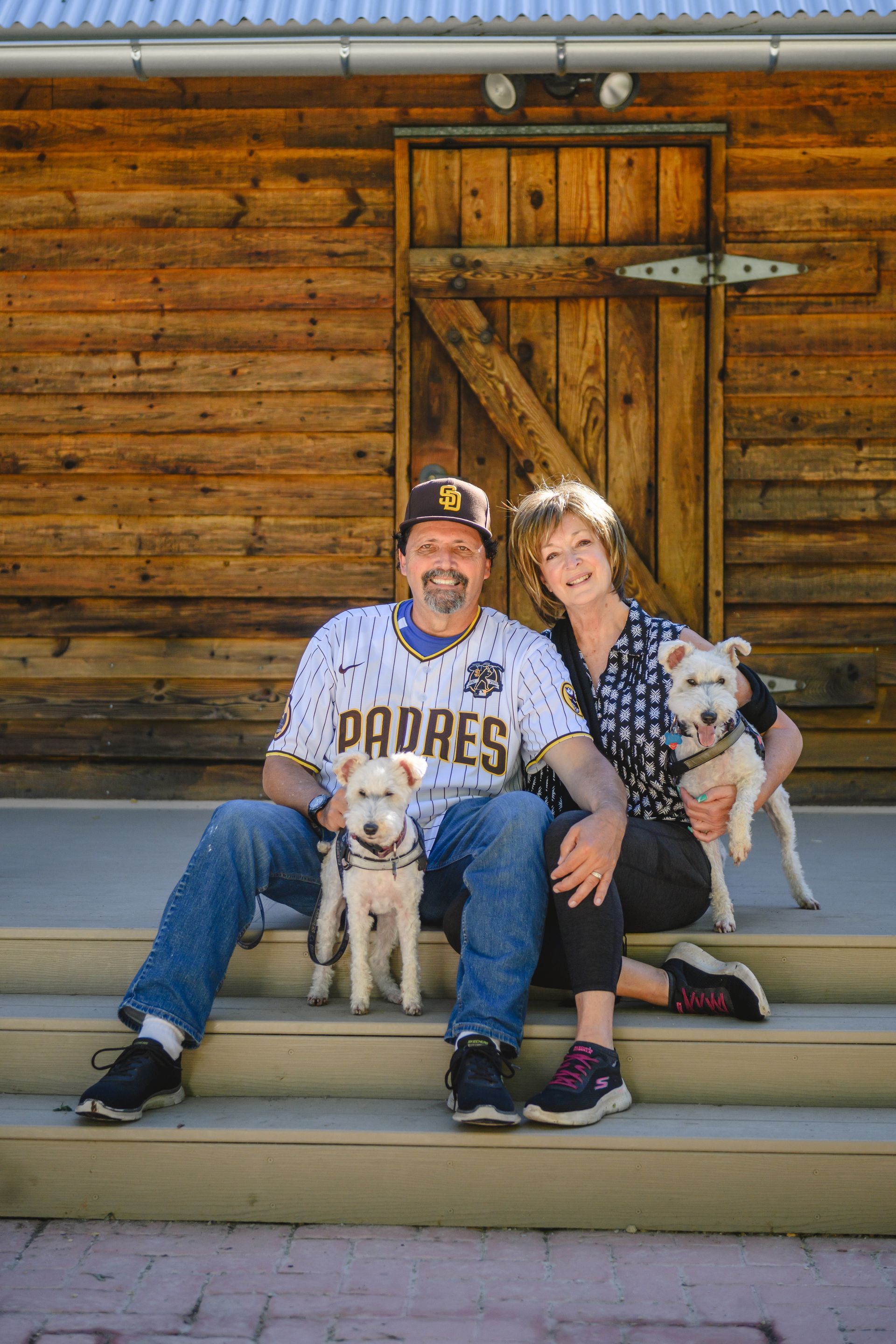 A couple sits on wooden porch steps in front of a log building, each holding a small white terrier-type dog.