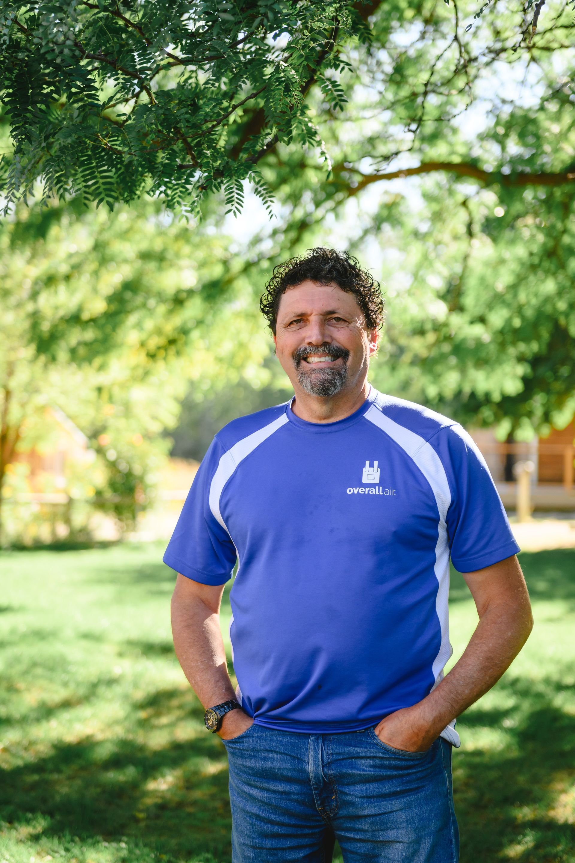 A person in a blue athletic shirt with white side panels and jeans stands outside on a grassy lawn under a tree.