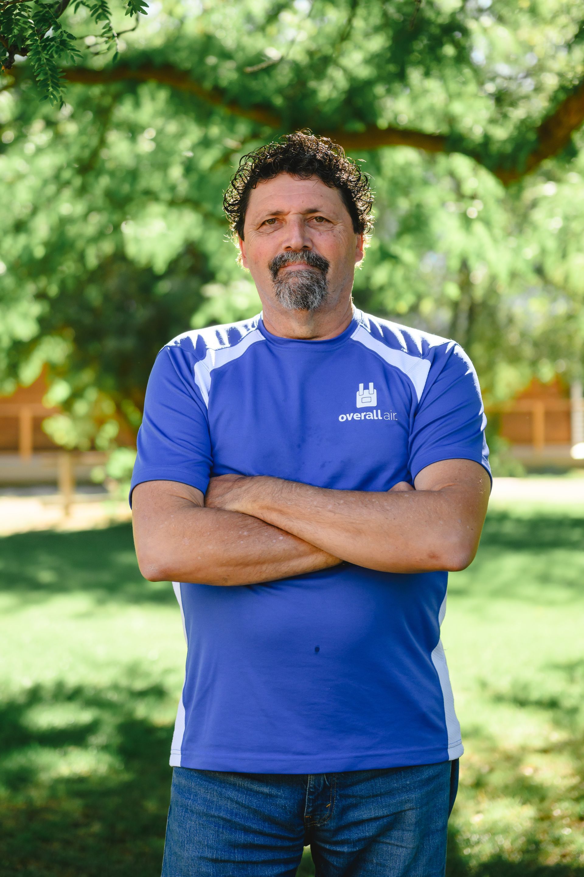 A man with dark curly hair and a beard, wearing a blue t-shirt with arms crossed, stands outdoors in a grassy park.