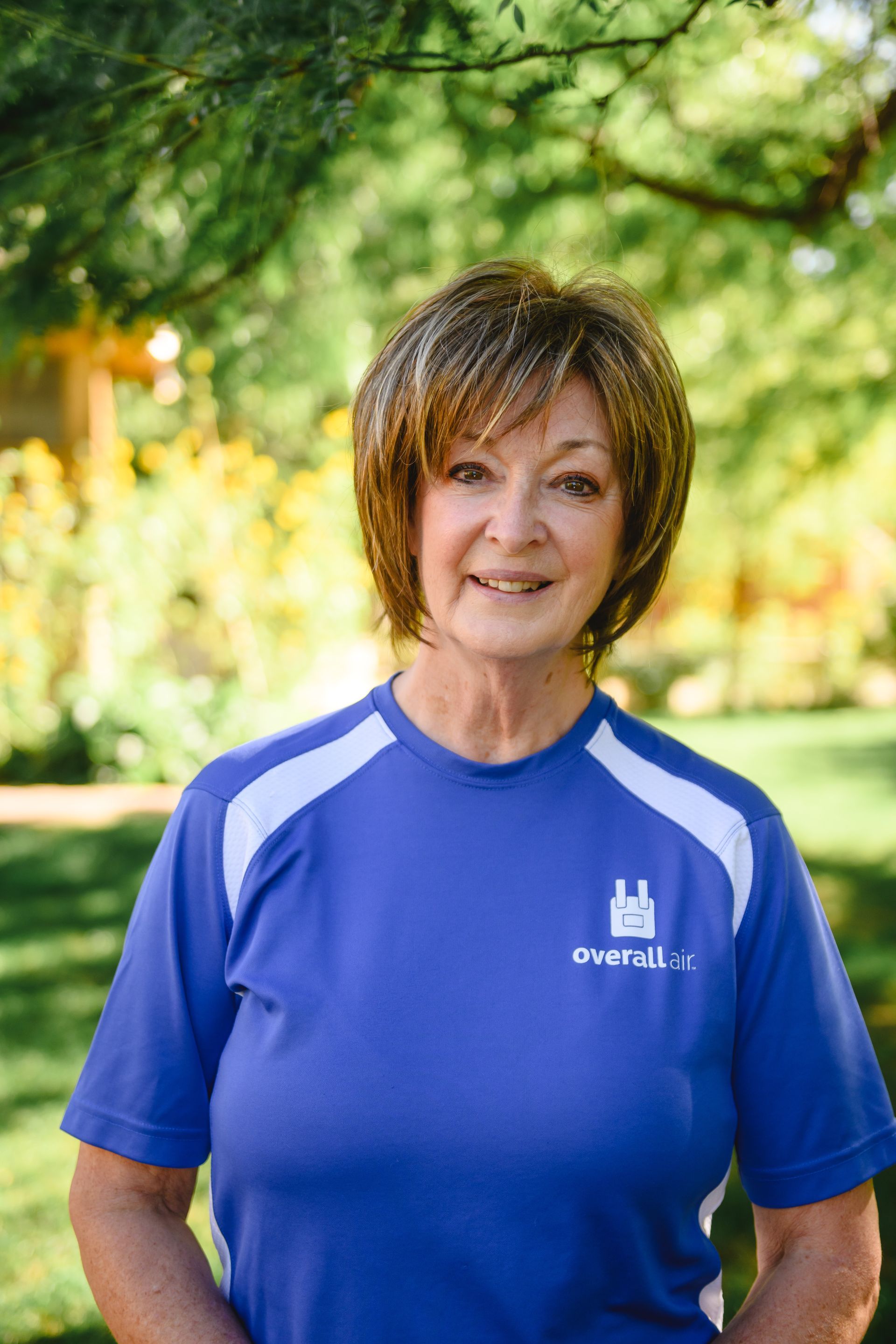 A woman with brown hair smiles, wearing a blue short-sleeved athletic shirt with white shoulder panels in a garden.