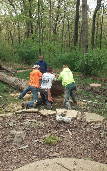 Four men lifting a large stone outdoors; forest background.
