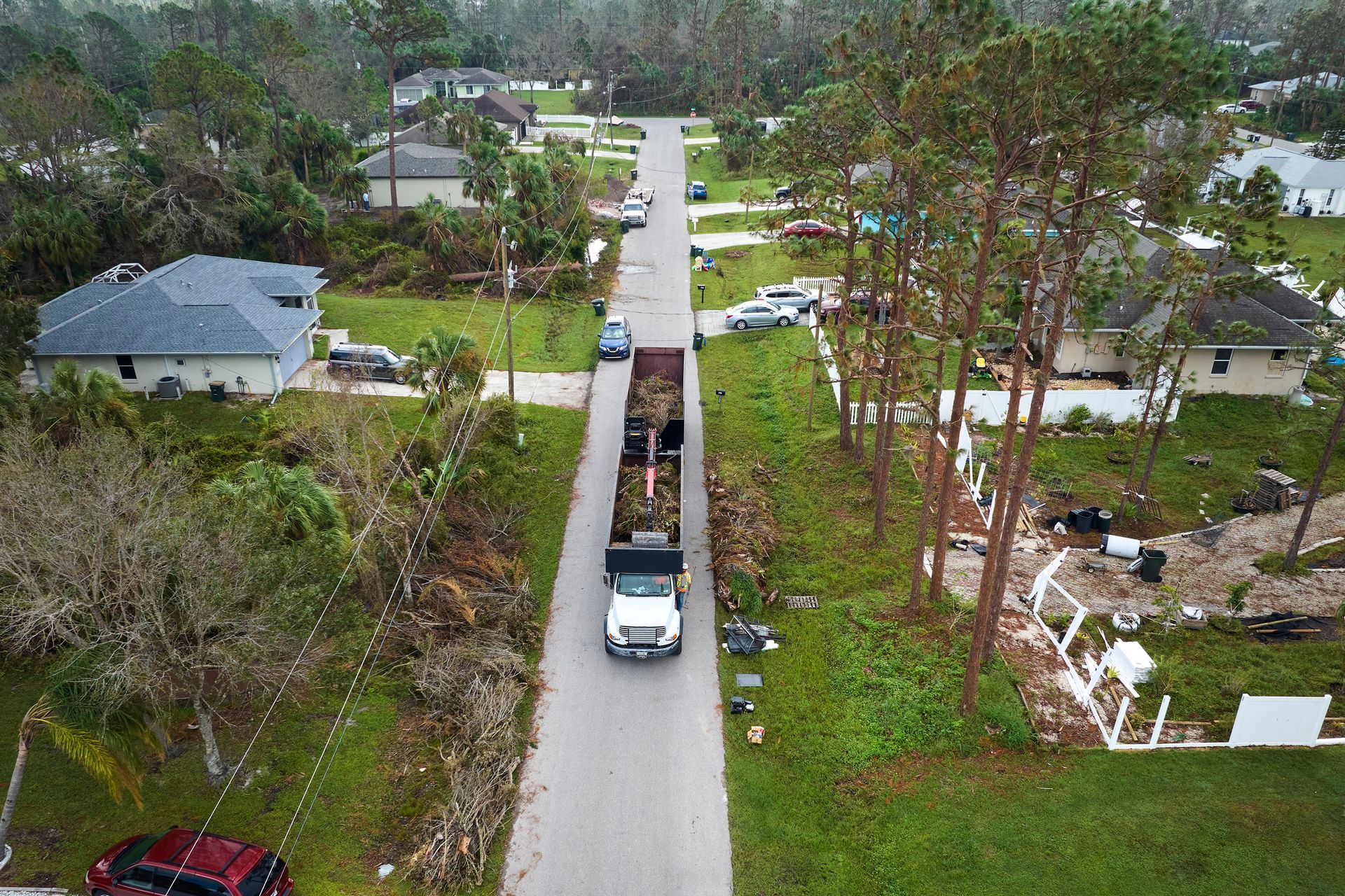 A truck collects debris from a residential street after a storm, with damaged trees and houses visible.