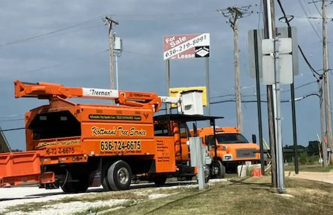 Two white utility trucks with extended booms parked on a tree-lined street, orange traffic cones set out.