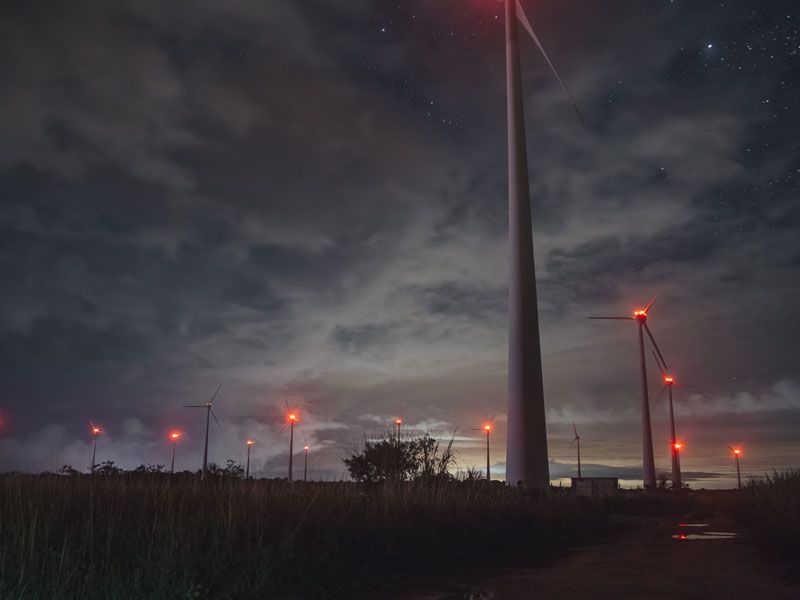 Turbinas eólicas ao entardecer sob um céu nublado com luzes vermelhas de advertência.
