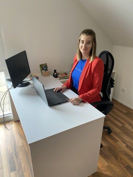 Eine Frau in einer roten Jacke sitzt mit einem Laptop an einem Schreibtisch.