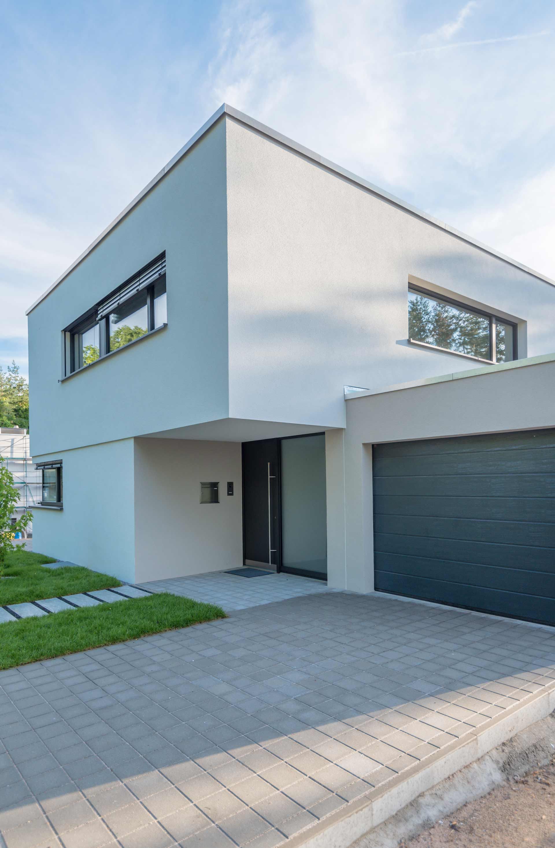 Modern two-story house with light gray facade, dark garage door, and a paved driveway.