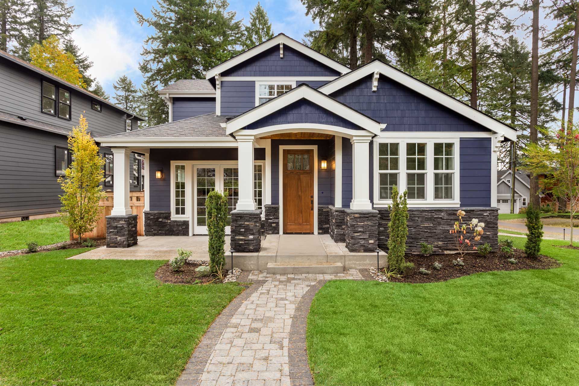 Blue house with white trim, brown door, stone accents, and green lawn; walkway leads to porch.