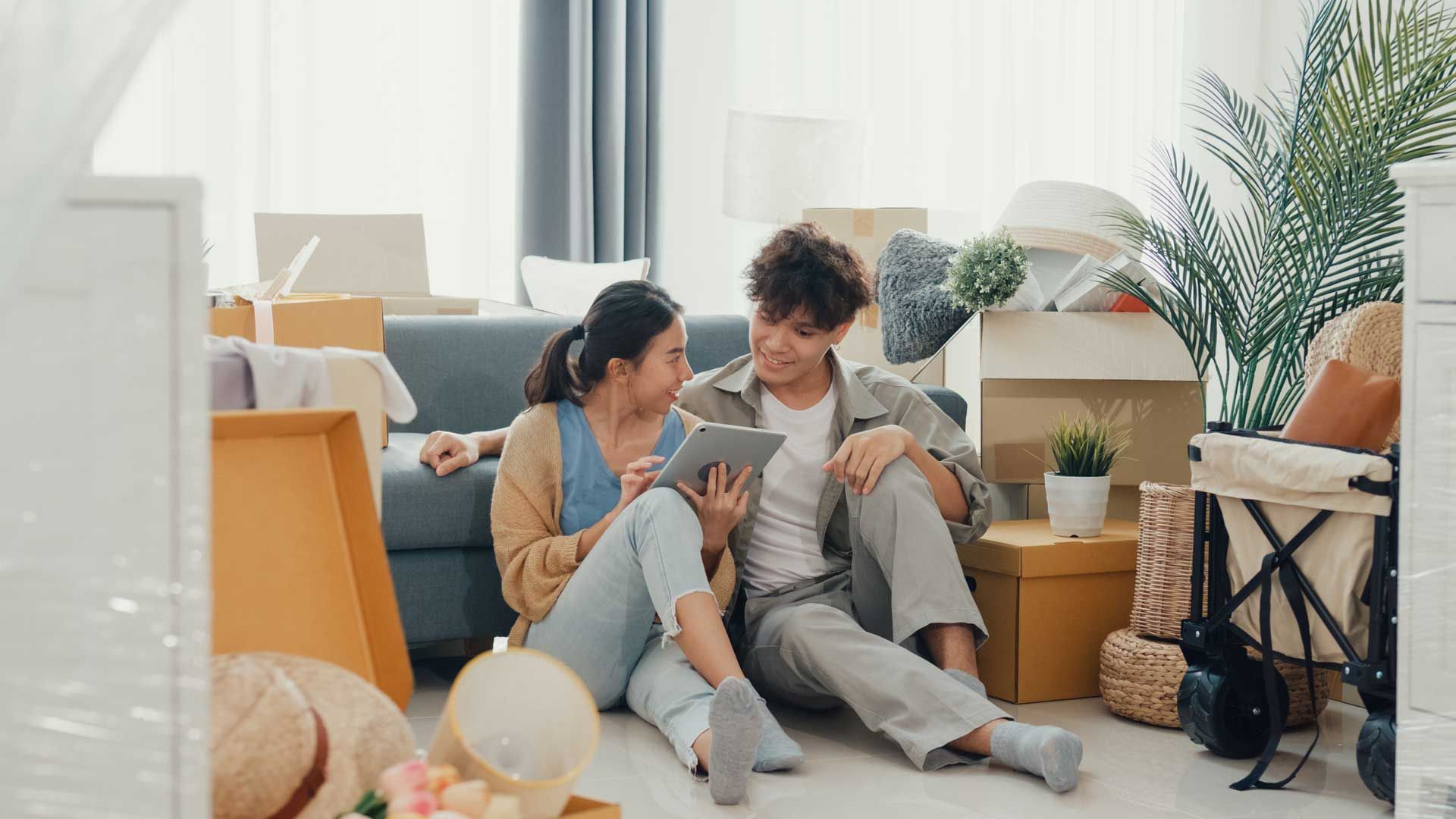 Young couple sits on floor surrounded by moving boxes, looking at a tablet, smiling. Living room.