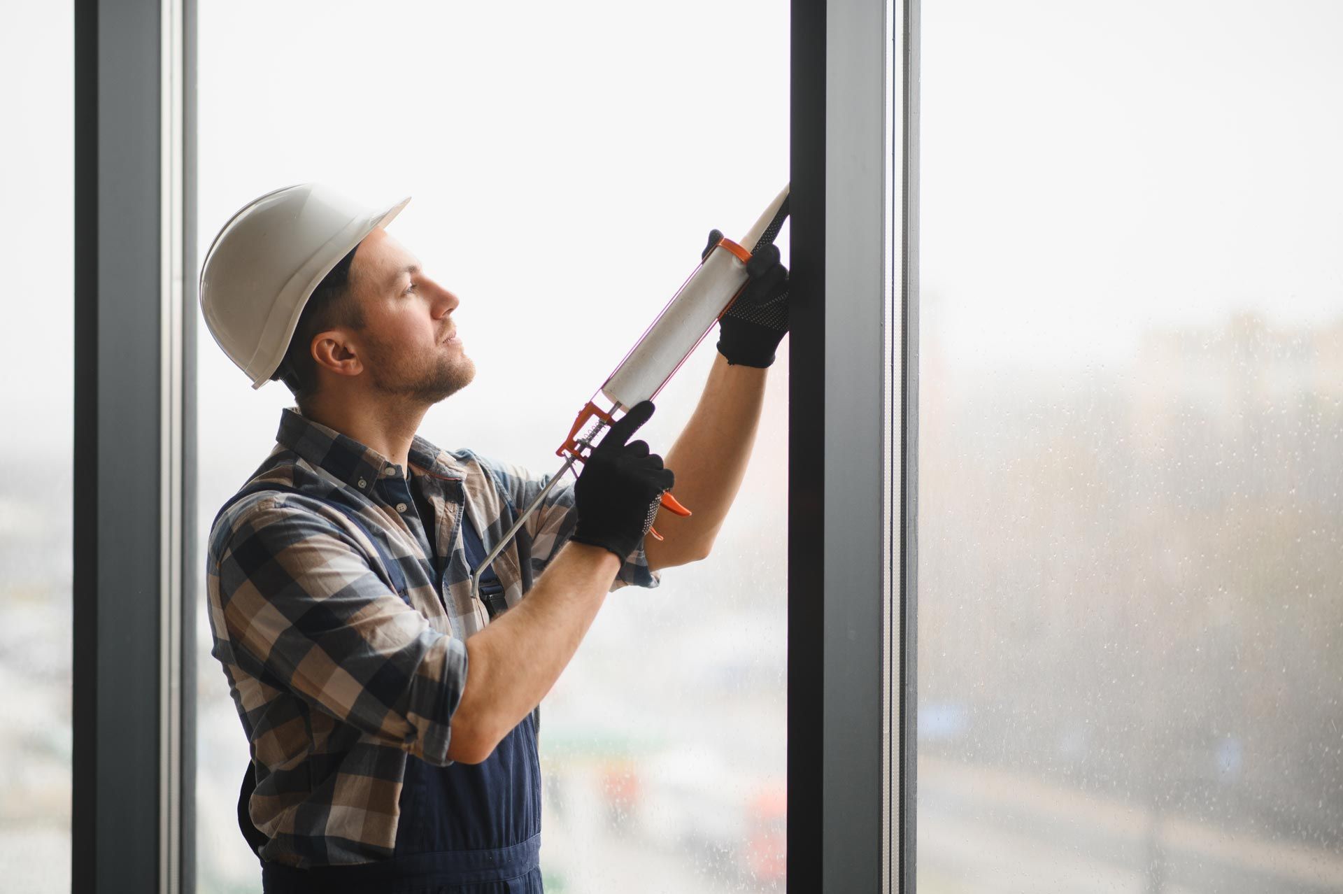 Construction worker wearing a hard hat and gloves applying sealant to a window frame.