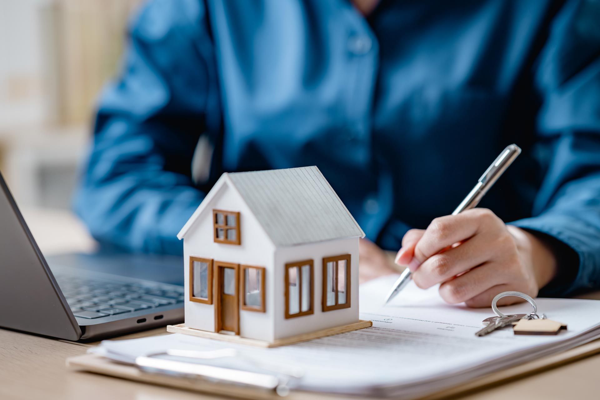 Person signing a document, miniature house, laptop, and keys on a desk.