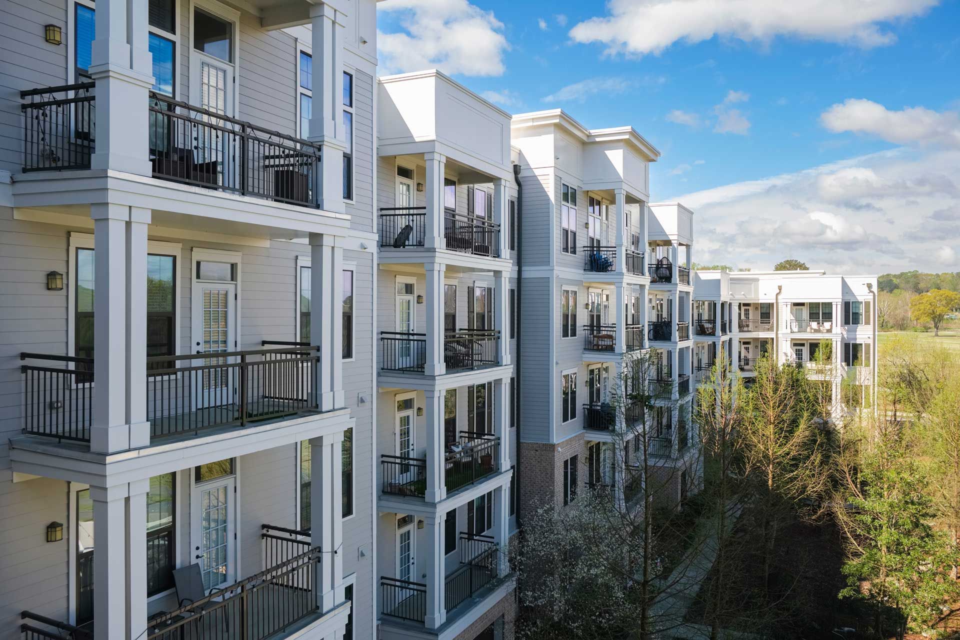 Multi-story apartment building with balconies, set against a blue sky with trees.