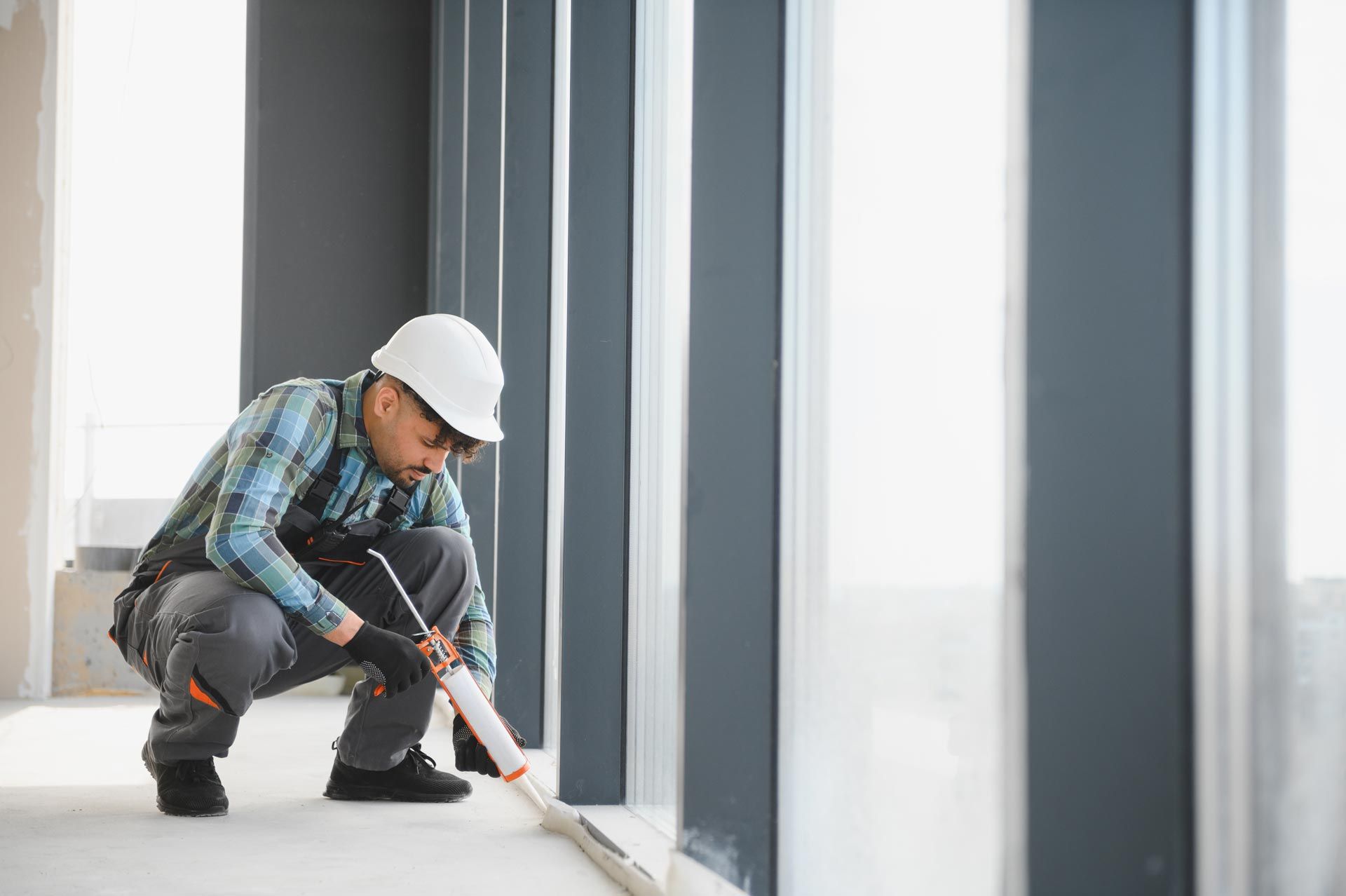 Construction worker caulking a window. Gray, white, and blue colors; indoor setting.