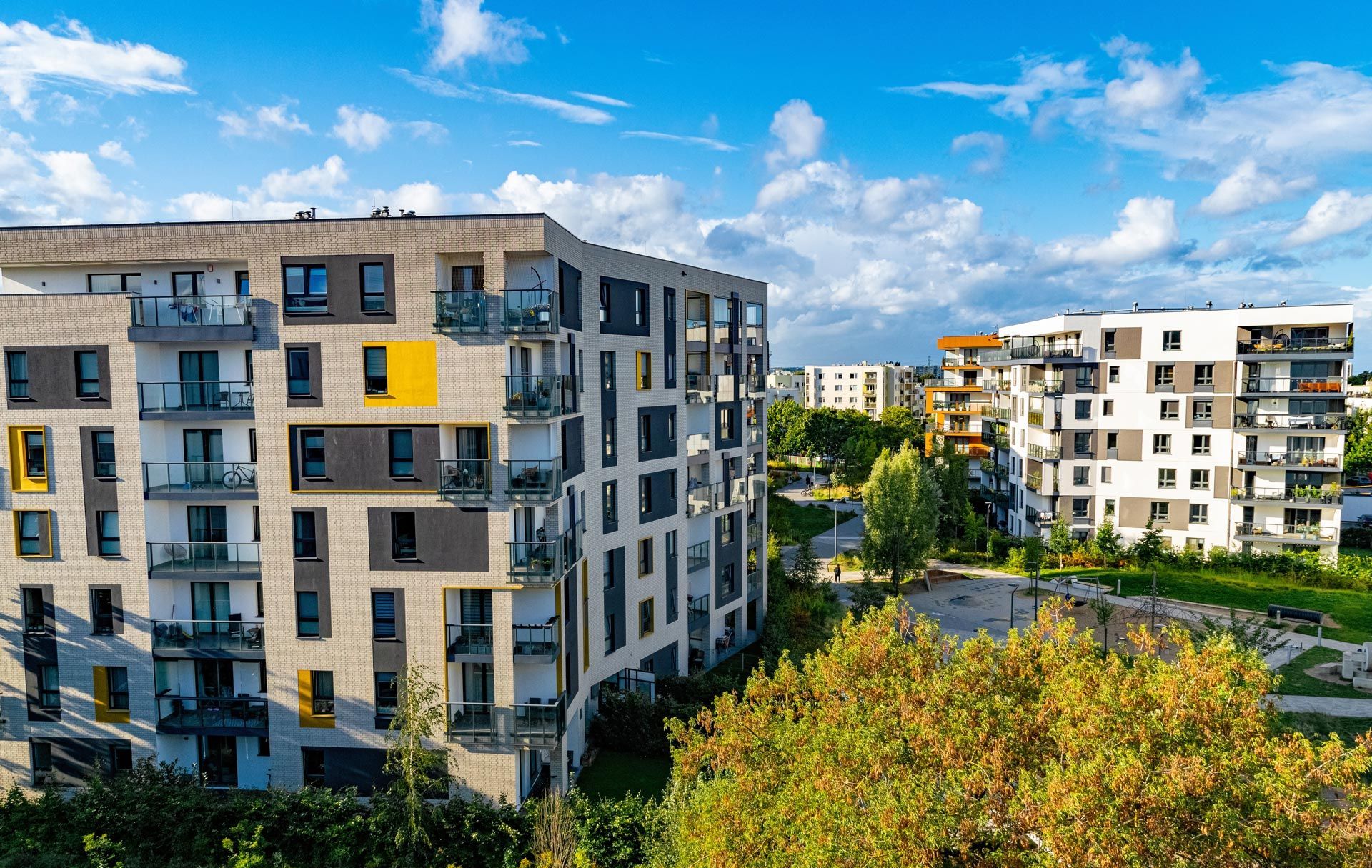 Modern apartment buildings with balconies against a blue sky, trees in foreground.