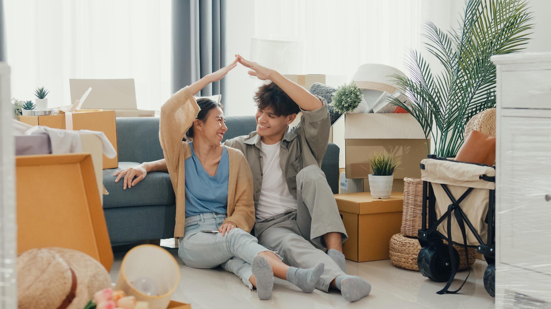 Couple sitting on floor, hands forming roof over their heads, surrounded by moving boxes in a living room.