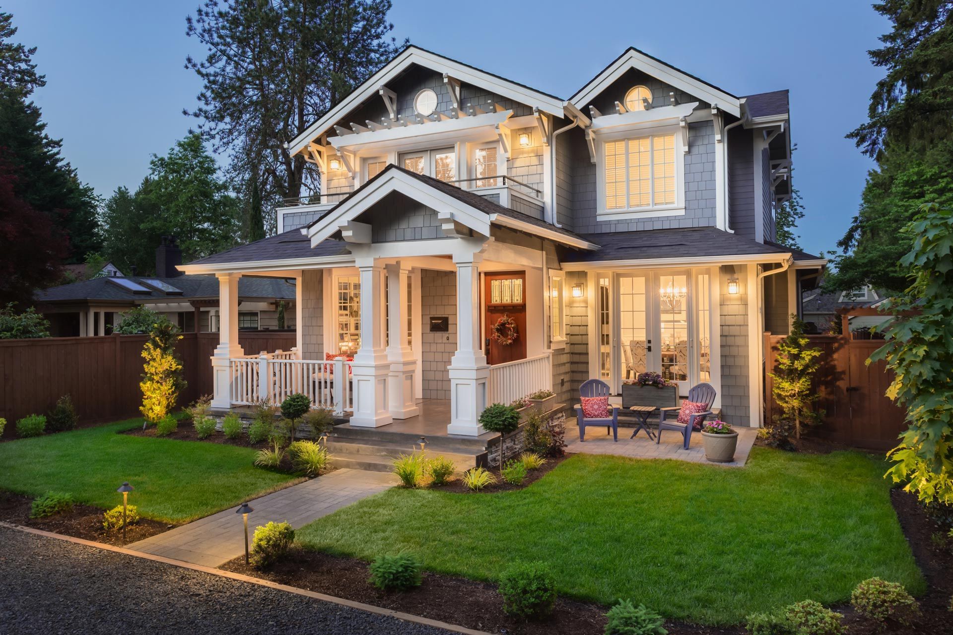 Two-story gray house with porch, lit windows, and manicured lawn. Dusk setting.