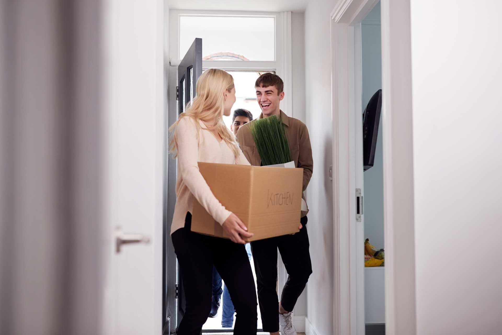 People carrying boxes and a plant into a new home doorway. Bright hallway.