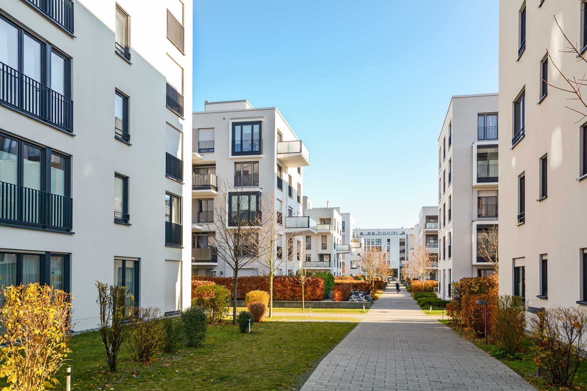 White apartment buildings with square windows and black railings on a sunny day. A walkway and green space lie between them.