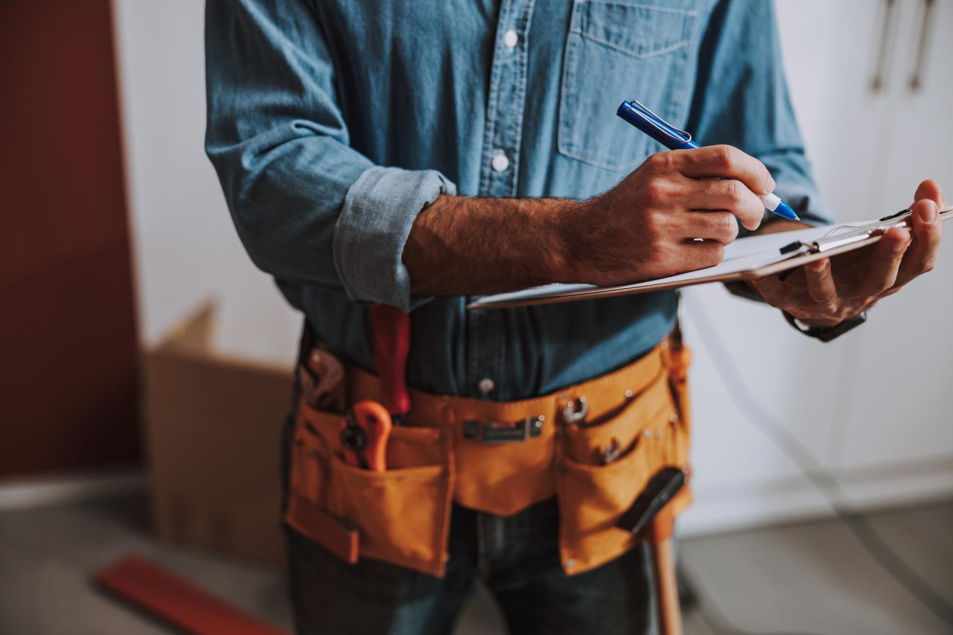 Person in denim shirt with tool belt writing on clipboard.